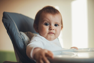 Close-up portrait of a hungry baby looking desperately at the camera sitting in the high-chair. Positive person. Baby care.
