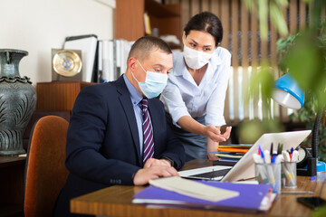 Fototapeta na wymiar Two business colleagues wearing protective face masks focused on working with laptop and papers in modern office. Necessary precautions during coronavirus pandemic..