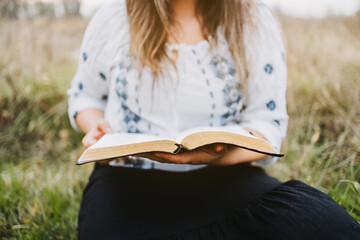Unrecognizable woman sitting on the grass holding and reading an open Bible. Selective focus.