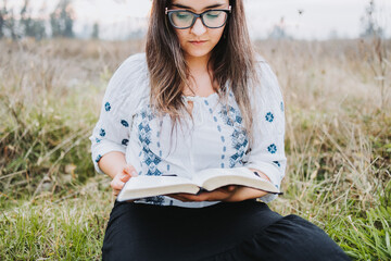 Woman with glasses sitting outdoor on the grass holding and reading an open Bible. Selective focus.
