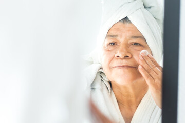 facial care Senior latin woman smiling applying face cream in front of mirror