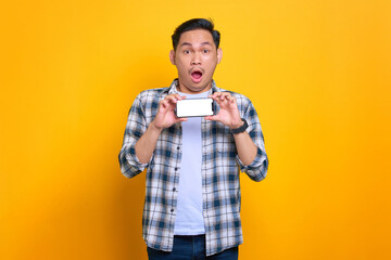 Shocked young Asian man in plaid shirt showing mobile phone blank screen recommending app isolated on yellow background