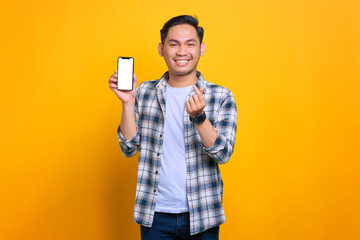 Cheerful young Asian man in plaid shirt holding mobile phone with blank screen, showing korean heart gesture isolated on yellow background