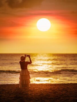 Asian woman come to travel to relax at the sea on vacation using a smartphone takes pictures of the sunset silhouette alone in the evening at Khao Lak Beach, Phang Nga, Thailand.