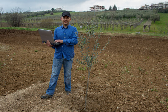 Photo of a smiling farmer with a laptop in his hand after planting an olive tree in a field.