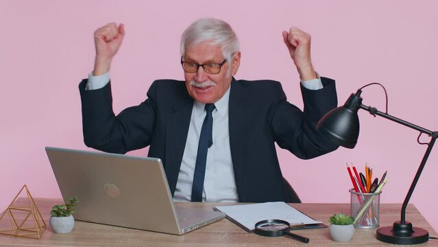 Excited amazed senior businessman in suit looking shocked surprised wow showing hands brain explosion head, sits at workplace desk at office. Elderly man isolated indoor on pink studio wall background