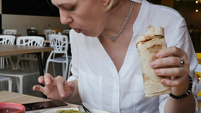 a caucasian woman in a white shirt in a cafe is having lunch at a table holding a shawarma in one hand and using a smartphone with the other hand continues to work online. slow motion