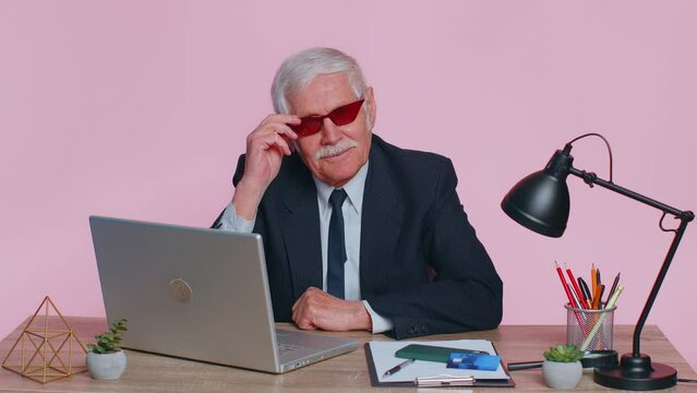 Senior funny business man in sunglasses using laptop computer notebook, raises thumbs up agrees with something or gives positive reply recommends advertisement likes good sits at desk at pink office