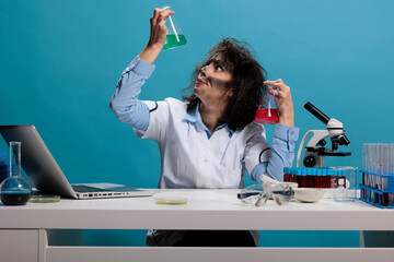 Potrait of crazy silly chemist analyzing glass jars filled with liquid experiment serums while sitting at desk. Foolish goofy looking maniac scientist having beakers filled with chemical compounds.