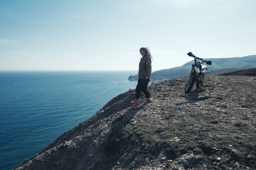 Woman rider traveling on enduro motorcycle, enjoying view from mountain cliff, beautiful sea shore...