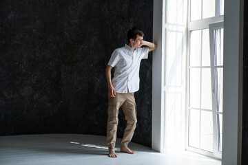 Fototapeta na wymiar Young pensive man wearing white shirt standing near big window looking outside