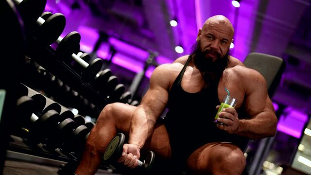 A bearded bodybuilder shakes his biceps with a dumbbell and drinks fresh orange juice, balanced fitness nutrition, workout