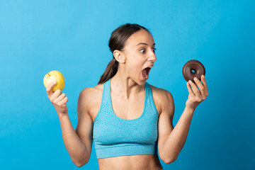 Surprised sportswoman looking at chocolate donut and holding apple isolated on blue background....