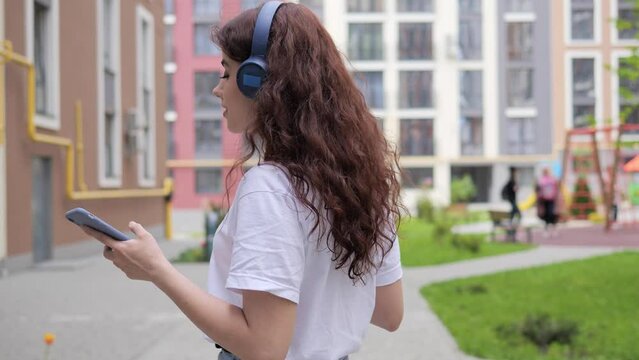 Attractive young curly hair woman with coffee listening to music in wireless headphone using mobile smartphone new app in urban street turning around looking posing to camera walking in the city.