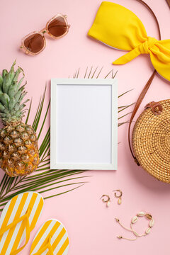 Top view vertical photo of white frame pineapple round rattan bag yellow swimsuit bodice slippers shell bracelet earrings sunglasses and palm leaves on isolated pastel pink background with copyspace