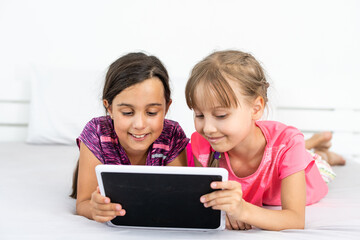 Two cute little girls are using a digital tablet and smiling while lying on bed in children's room