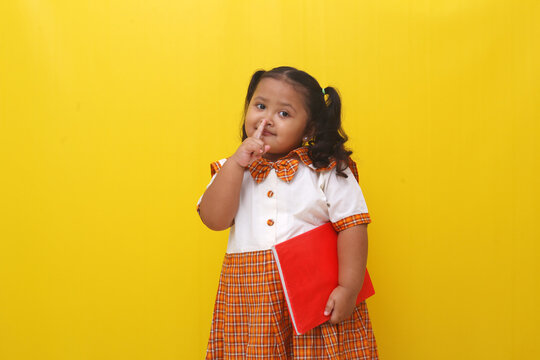 Asian little schoolgirl standing while holding a book with silence finger gesture.