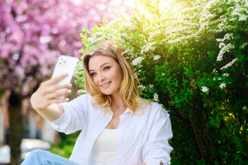 Beautiful blonde hair girl in a white shirt holds a phone and takes a selfie in the park. Sunshine