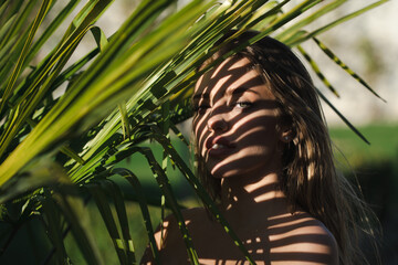 Woman hiding behind the palm leaves. Beautiful young woman face with shadows of tropical palm leaf....