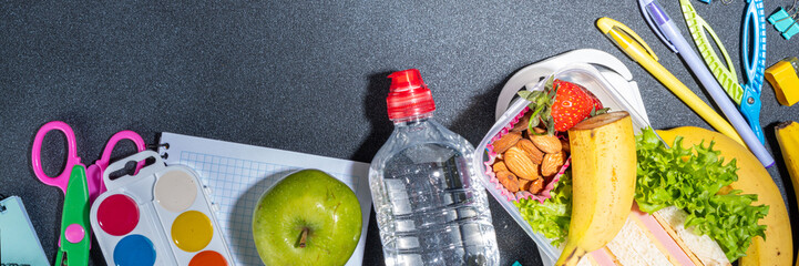 Healthy school lunch box: sandwich, vegetables, fruit, nuts and yogurt with school kids supplies, accessories and backpack on black background flatlay copy space. Back to school concept