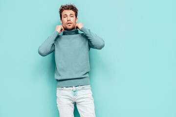 Portrait of handsome confident  model. Sexy stylish man dressed in  sweater and jeans. Fashion hipster male with curly hairstyle posing near blue wall in studio. Isolated