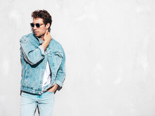 Obraz premium Portrait of handsome confident stylish hipster lambersexual model with curly hairstyle. Sexy man dressed in jeans jacket. Fashion male isolated on grey wall in studio. In sunglasses