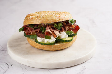 A bagel sandwich with pastrami, cucumber slices, watercress salad and ricotta on marble background