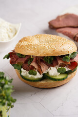 A bagel sandwich with pastrami, cucumber slices, watercress salad and ricotta on marble background