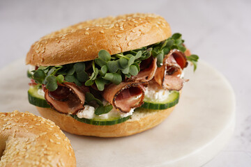A bagel sandwich with pastrami, cucumber slices, watercress salad and ricotta on round marble board