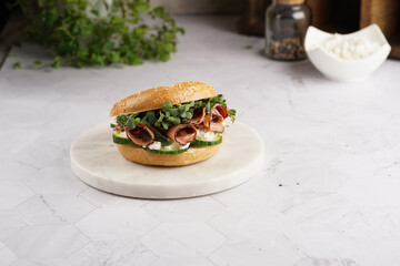 A bagel sandwich with pastrami, cucumber slices, watercress salad and ricotta on round marble board