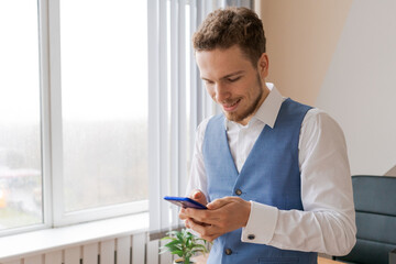 Naklejka na ściany i meble Young happy business man with beard smiling while reading his smartphone. Portrait smiling businessman reading message with smartphone in office. Man working at his desk in office standing by window