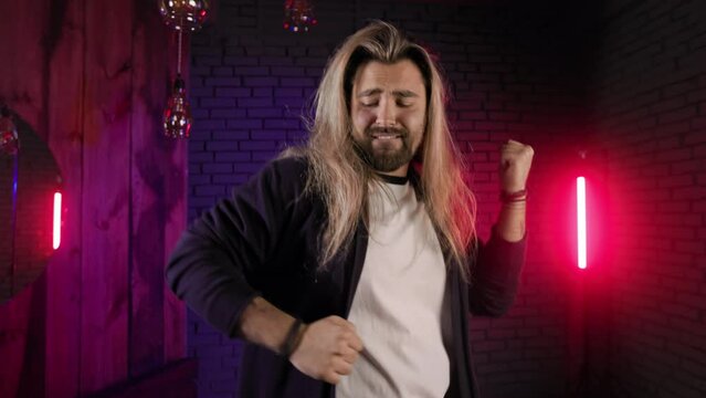 Joyful caucasian male person with long hair rocking alone in studio with neon light around. Bearded guy dancing rhythmically while enjoying his favorite style of music.