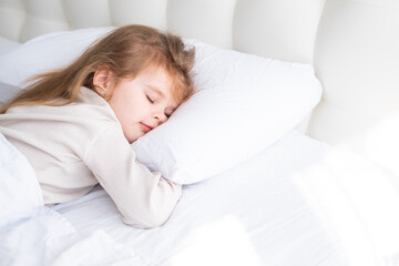 Fototapeta premium cute child girl with long hair in beige pyjamas sleeping on white bedding at home