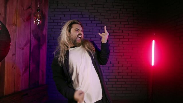 Caucasian young man with long hair playing air guitar in studio with neon light. Fun and pleasure of male hard rock musician.