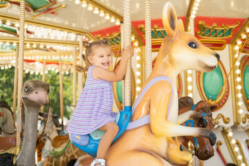 funny little kid girl in colorful dress rides on carousel in an amusement park in summer day