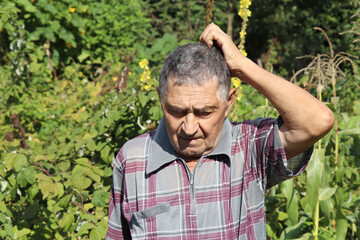 Portrait of elderly man scratches his head standing in summer garden. Concept of life in village,...