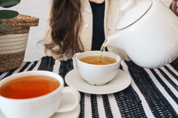Cropped view of young woman spending morning standing on kitchen, pouring traditional tea from transparent teapot into a glass cup. Female make hot weight loss drink with organic herbs