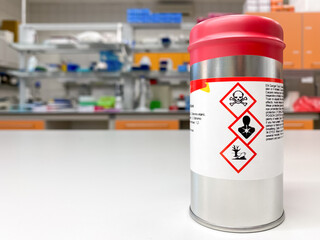 Can with extremely dangerous substance inside, labelled with symbols indicating that the content is toxic. Laboratory space is visible in the background. - obrazy, fototapety, plakaty