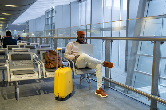 Pensive African American man sitting in airport lounge using laptop, wear headphones listening music. Passenger travelling with backpack and suitcase, online working on computer and waiting landing. 