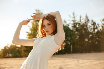 Confident redhead female model in white dress standing with raised hands in nature in sunset and looking at camera 