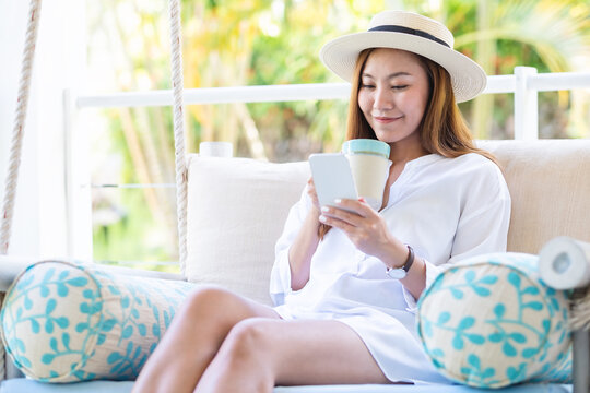 Portrait image of a beautiful asian woman holding and using mobile phone while drinking coffee and sitting on swing sofa