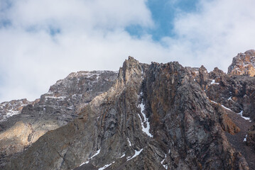 Atmospheric landscape with sharp pinnacle on sunlit high mountain range under clouds in blue sky. Beautiful alpine scenery with peaked mountain top in sunlight under cloudy sky at changeable weather.