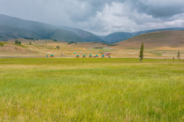 Scenic view from sunlit grassy steppe to small houses on background of green high mountain range in rainy clouds. Dramatic landscape with meadow and small lodges in mountains at changeable weather.