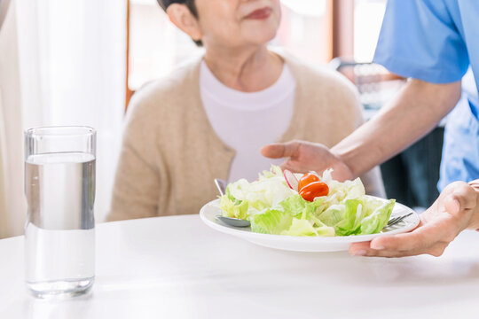 Close up hands of caregiver wearing medical scrubs caring to senior Asian woman by serving a vegetarian salad eating meals at home. Caregiver visit at home. Home health care and nursing home concept.