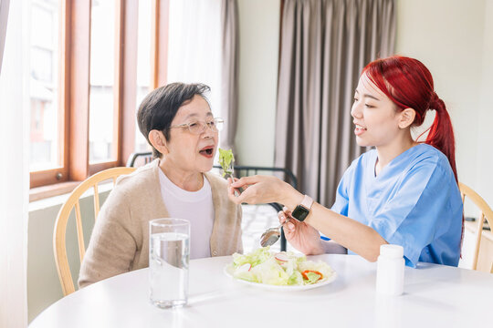 Asian woman caregiver wearing medical scrub takes care of senior Asian woman by feeding vegetarian salad to eat at home. caregivers visit home Home health care concept and nursing home.
