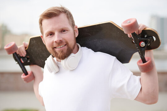 Portrait of happy young man with beard holding skateboard on shoulders outdoors