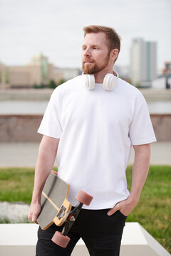 Confident hipster young bearded man with headphones around neck holding skateboard outdoors and looking into distance