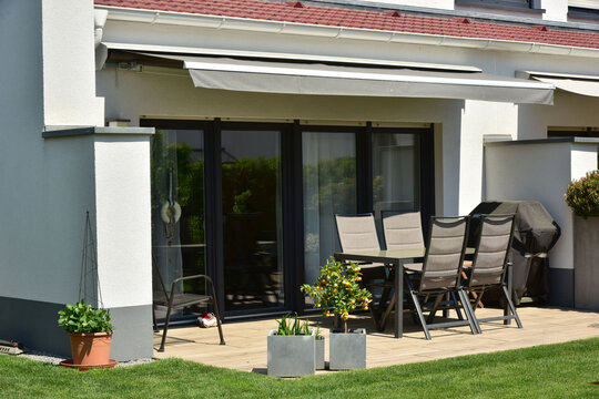 Terrasse und Gartenbereich mit Ziergeh&ouml;lz und Balkon und Ziegel-Vordach hinter einem Reihenhaus mit Essbereich, Gartenm&ouml;beln und Elektro-Markise als Sonnenschutz