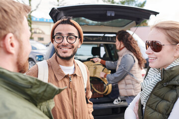 Group of positive young multi-ethnic planning travel route while girl packing bags into car trunk in background