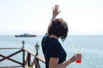 The girl is waving to a ship sailing in the distance while standing on the shore with a glass of juice.